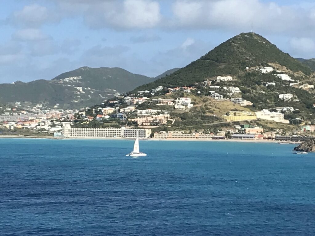 View of a tropical island coastline with turquoise water, hillside homes, and a sailboat crossing the harbor under a partly cloudy sky.