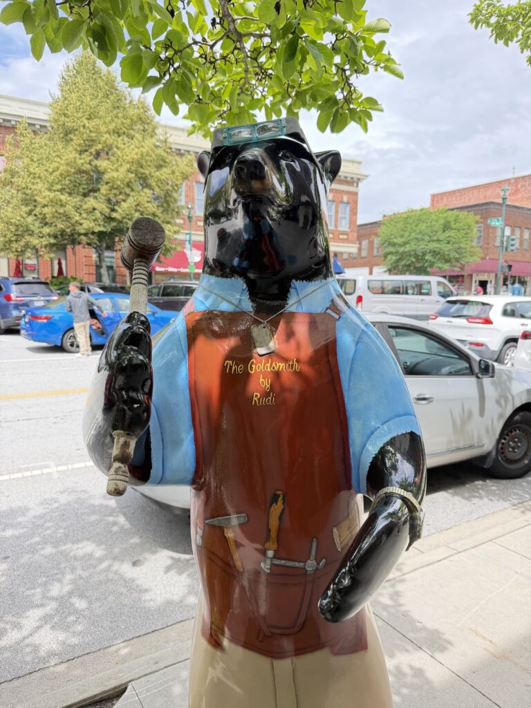 Decorative black bear sculpture dressed as a goldsmith stands along Main Street in downtown Hendersonville, North Carolina, holding a hammer and wearing a leather apron.