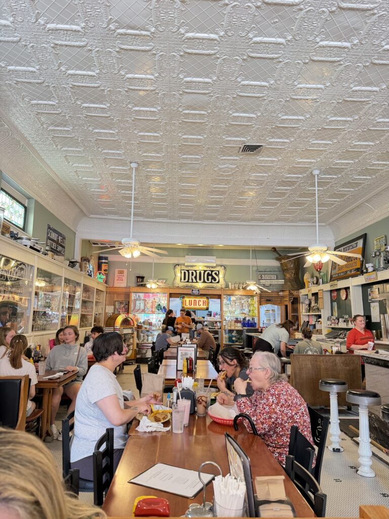Busy historic soda fountain and lunch counter inside a vintage drugstore in downtown Hendersonville, North Carolina, featuring ornate pressed-tin ceilings, antique décor, and diners enjoying meals.