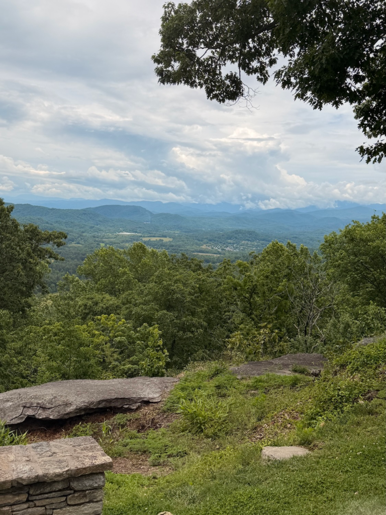 Panoramic mountain view from Jump Off Rock overlooking the Blue Ridge Mountains, with rolling forested hills, dramatic clouds, and a stone overlook wall in the foreground.