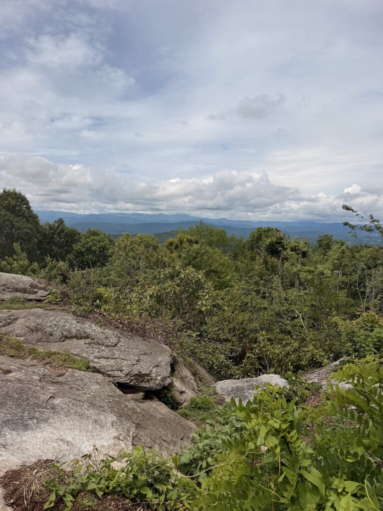 Mountain vista from Jump Off Rock in Laurel Park, North Carolina, featuring rocky outcroppings, dense forest, rolling Blue Ridge Mountains, and dramatic clouds stretching across the horizon.