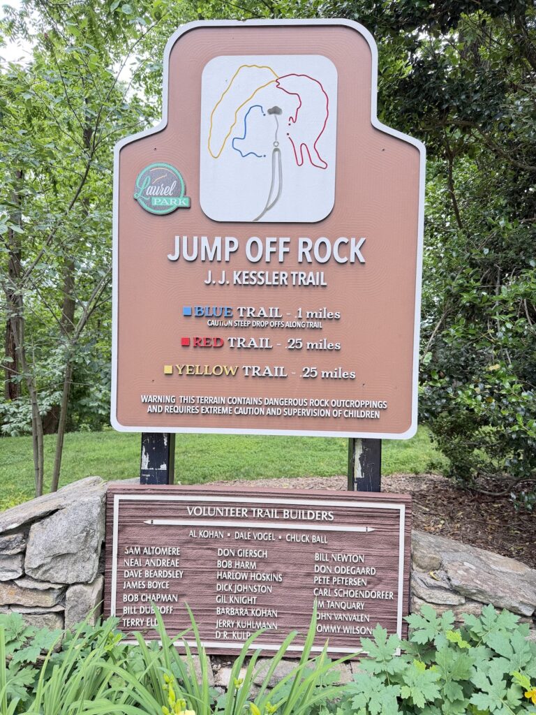Trailhead sign for the Jump Off Rock hiking trails in Laurel Park, North Carolina, showing trail distances, route colors, and safety information surrounded by lush forest.