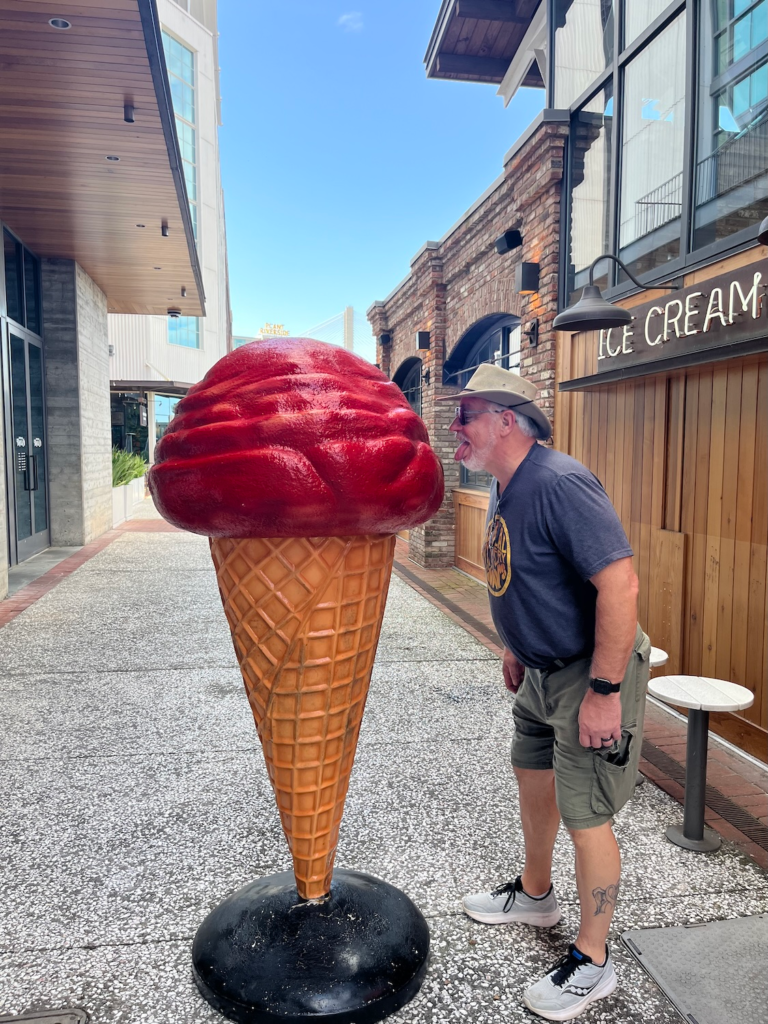 Man pretending to lick a giant red ice cream cone sculpture outdoors